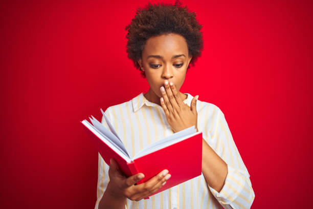 African american woman reading a book over red isolated background cover mouth with hand shocked with shame for mistake, expression of fear, scared in silence, secret concept